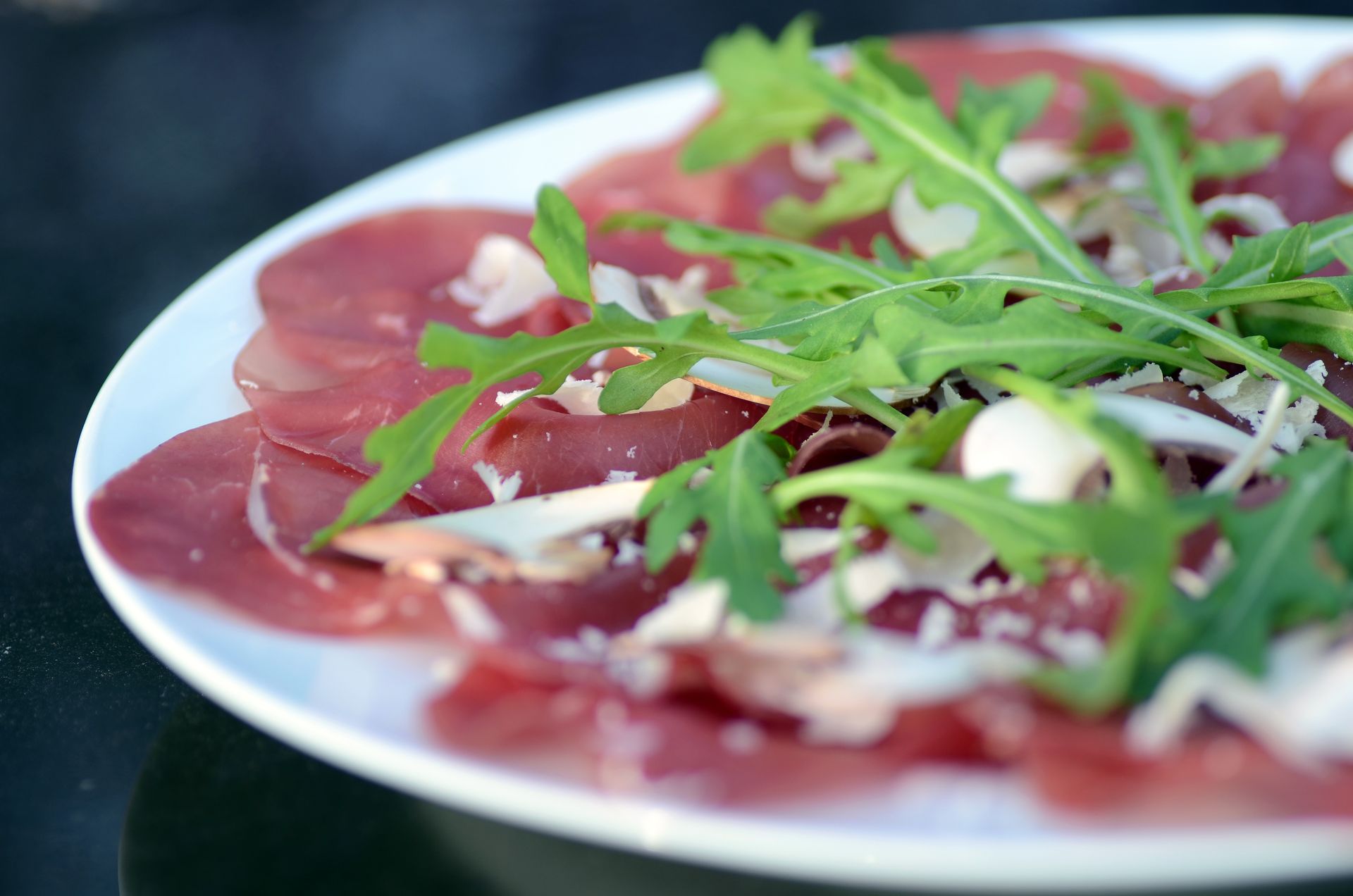 Dünn geschnittenes Wurstfleisch mit Rucola und gehobeltem Parmesan auf einem weißen Teller.