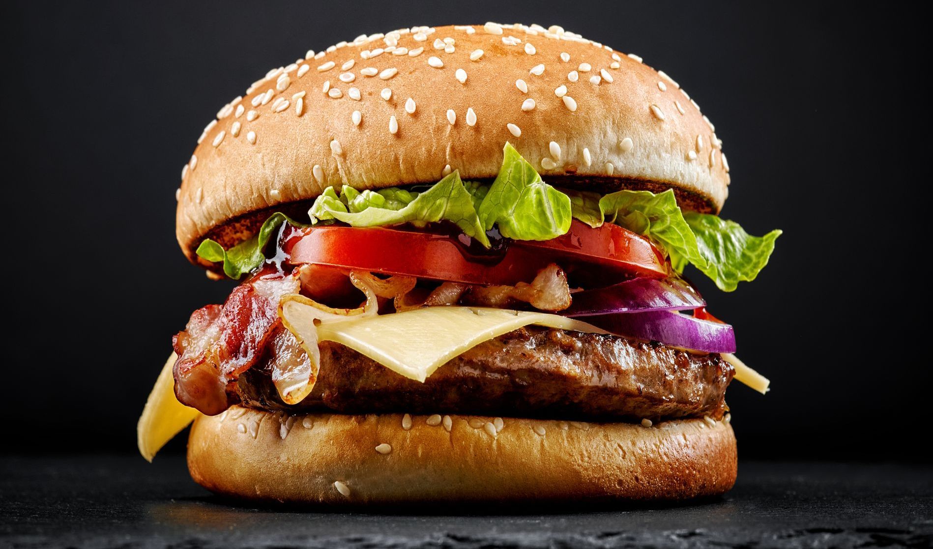 Burger mit Sesambrötchen, Speck, Käse, Tomaten, Salat und roten Zwiebeln auf dunklem Untergrund.