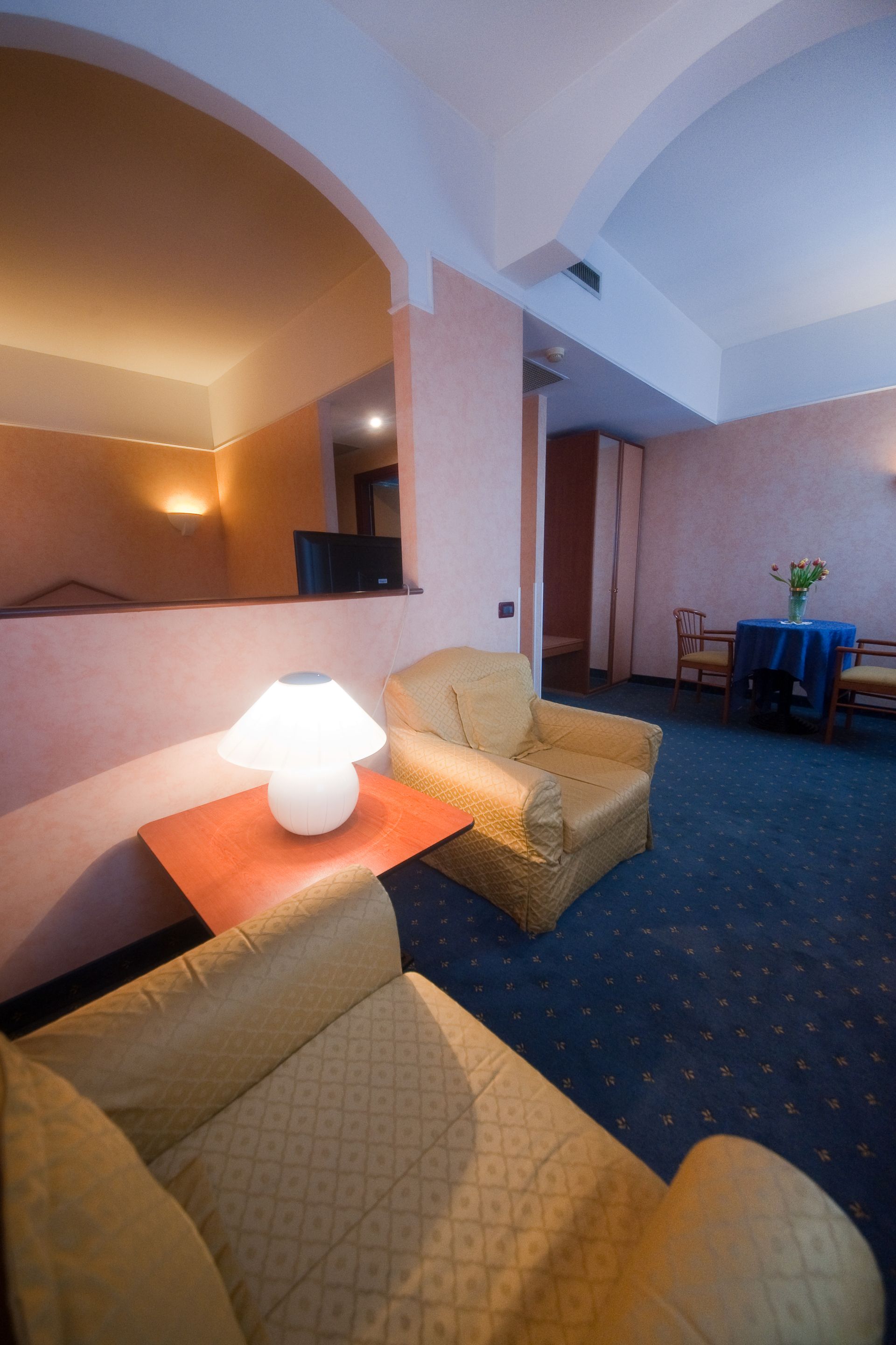 Interno della camera d'albergo: due poltrone gialle, tavolino con lampada, tappeto blu, arco.