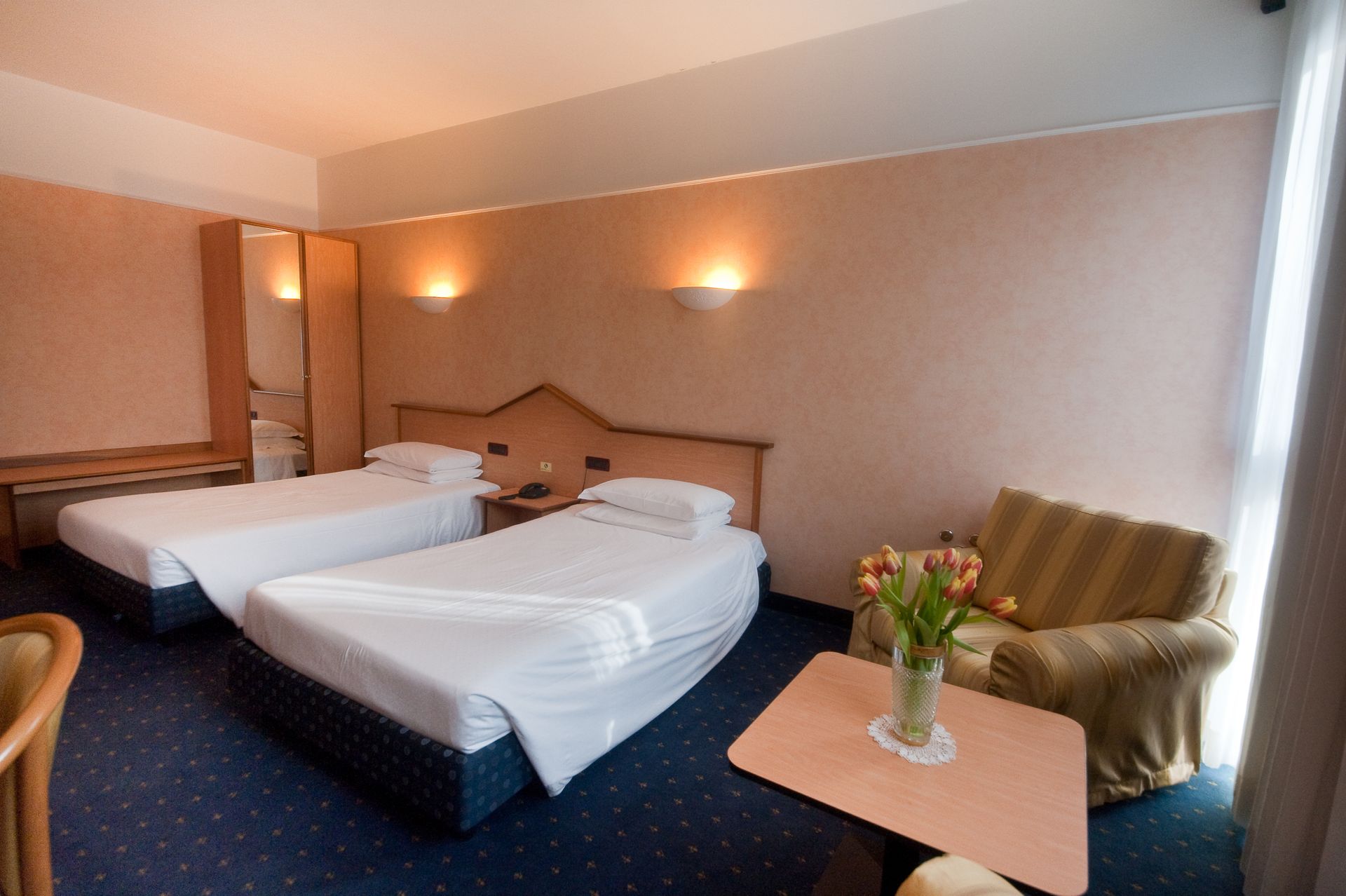 Camera d'albergo con due letti singoli, una poltrona e un tavolino con fiori. Moquette blu, pareti color pesca.