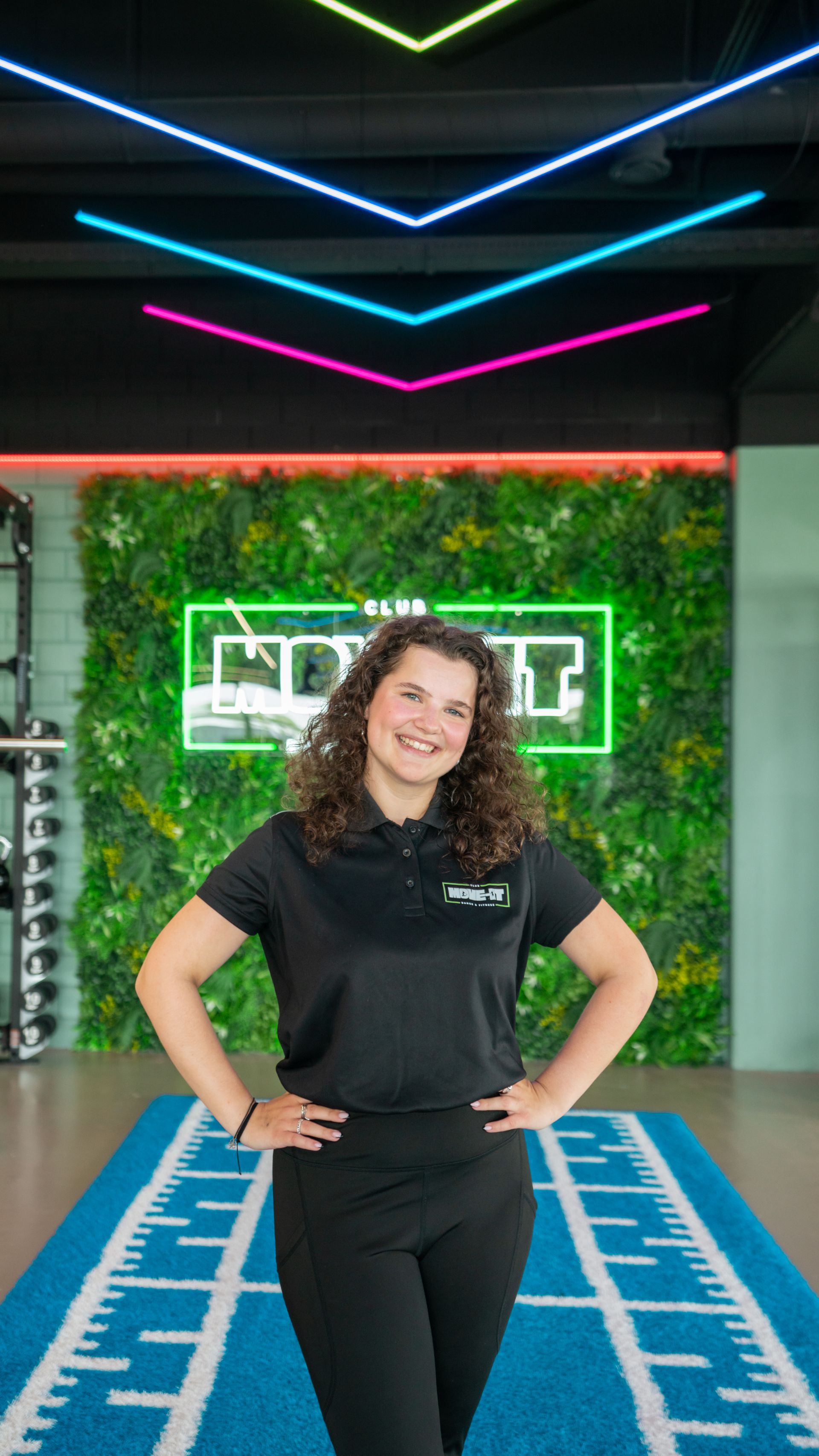 Een vrouw in een zwart shirt staat in een sportschool met haar handen in haar zij.