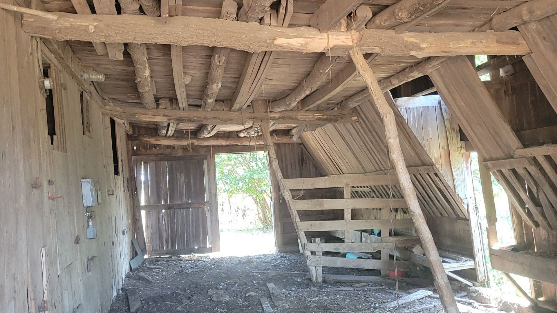 Interior of a dilapidated wooden barn with a staircase and open doorway. Sunlight streams in.
