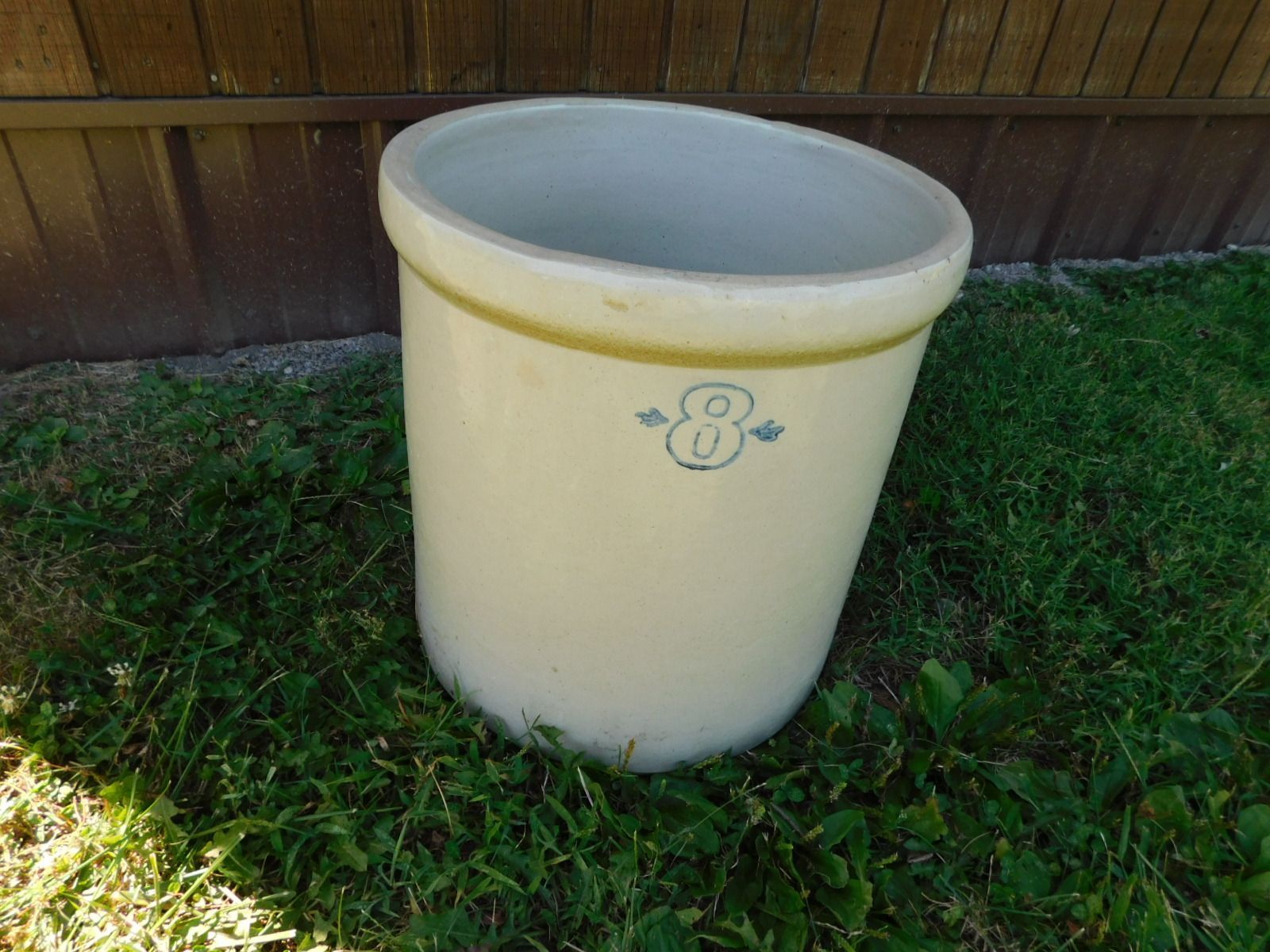 A large white crock is sitting in the grass in front of a wooden fence.