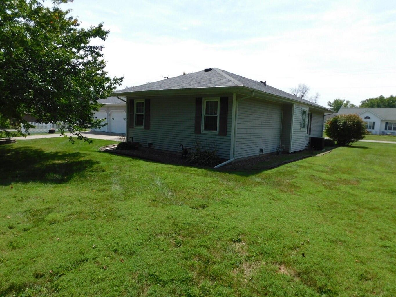 A one-story house with light siding and a dark roof sits on a grassy lawn on a sunny day.