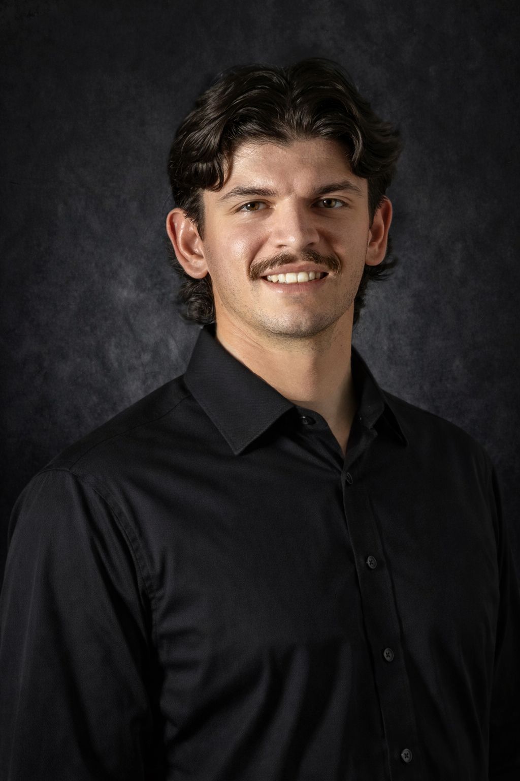 Man with wavy blonde hair wearing a gray polo shirt, looking at the camera against a dark backdrop.