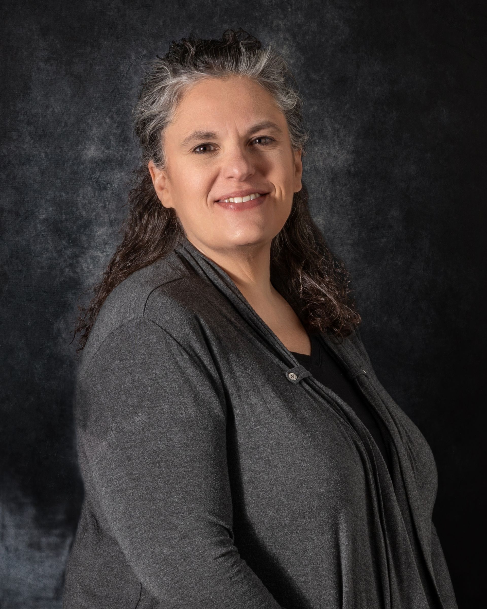 Man wearing glasses and camouflage hat smiles, gray collared shirt, dark background.