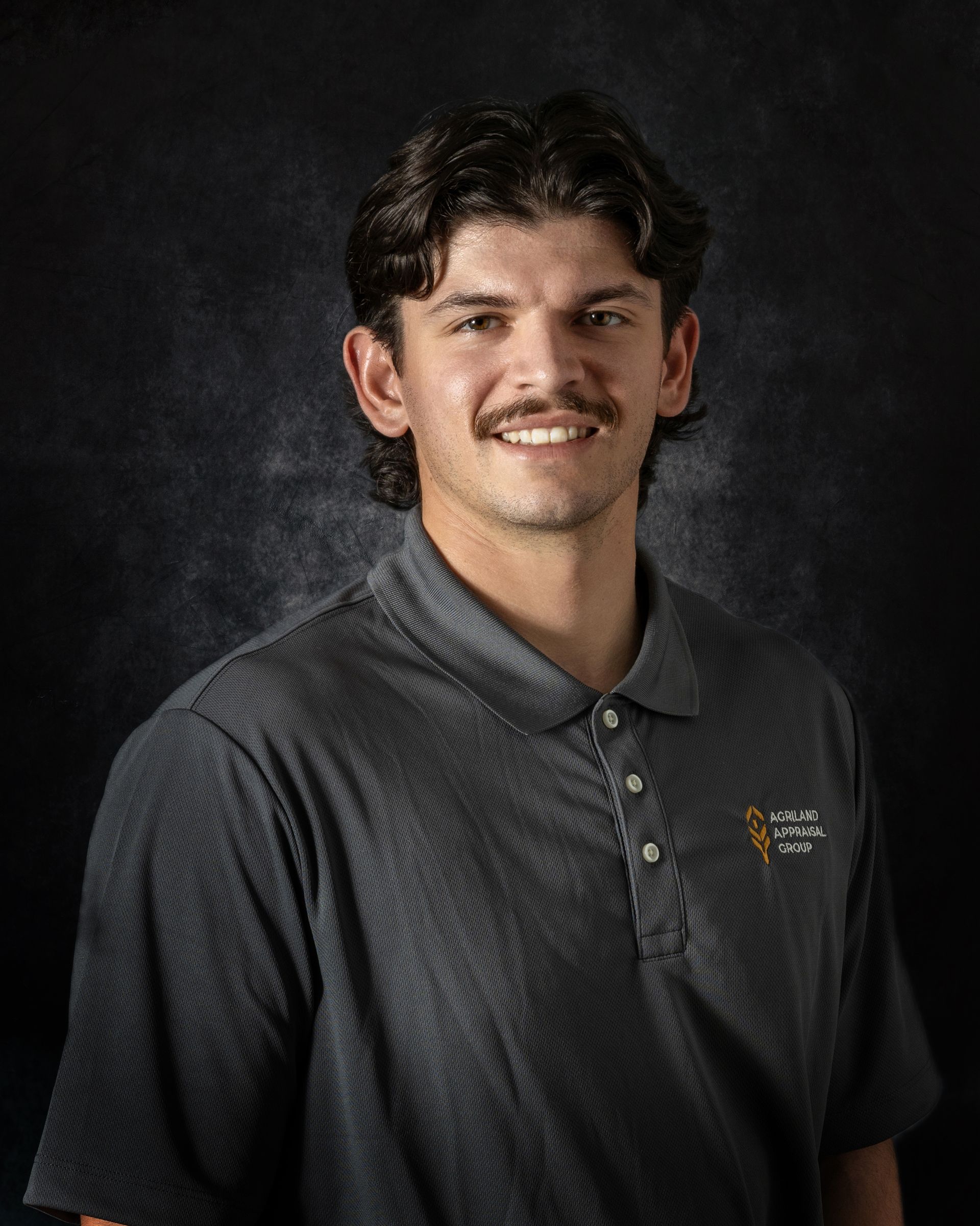 Man in gray polo shirt smiles, dark hair, thin mustache, against a dark backdrop.