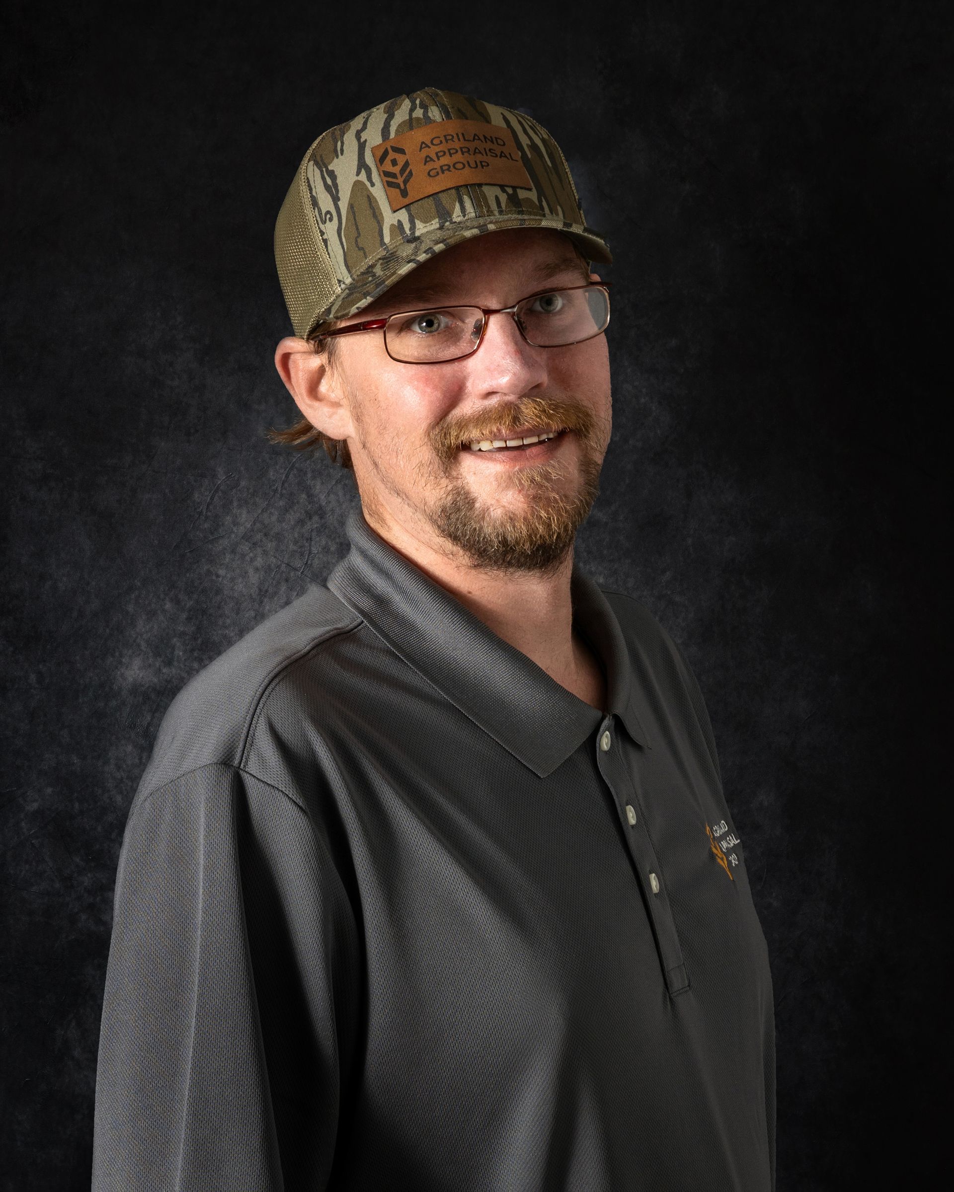 Man wearing glasses and camouflage hat smiles, gray collared shirt, dark background.