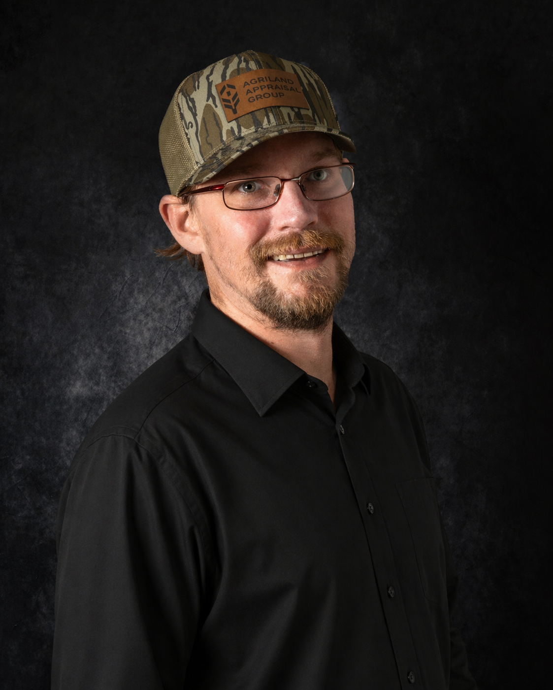 Man in camo hat and glasses, smiling, wearing a gray polo shirt. Dark background.