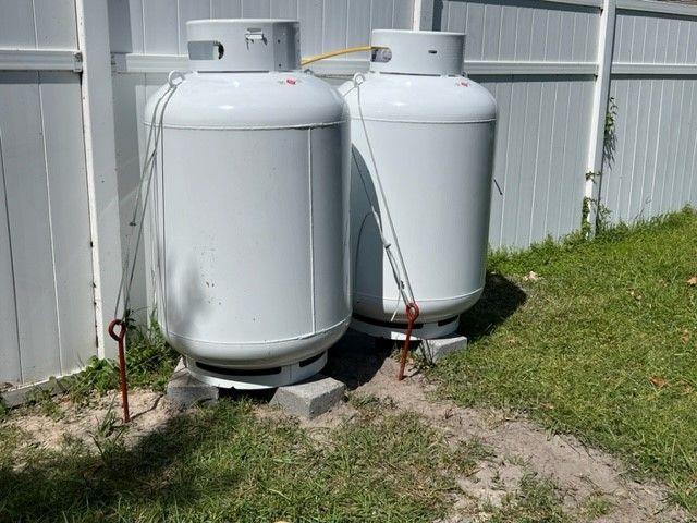 Two white propane tanks outdoors, secured with stakes, next to a white fence on a grassy area.