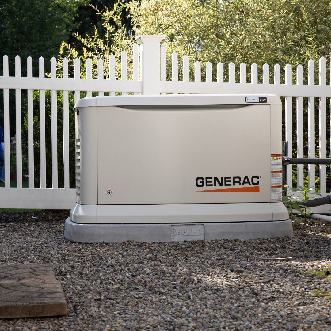 A Generac home backup generator, beige and gray, sits on gravel near a white picket fence.