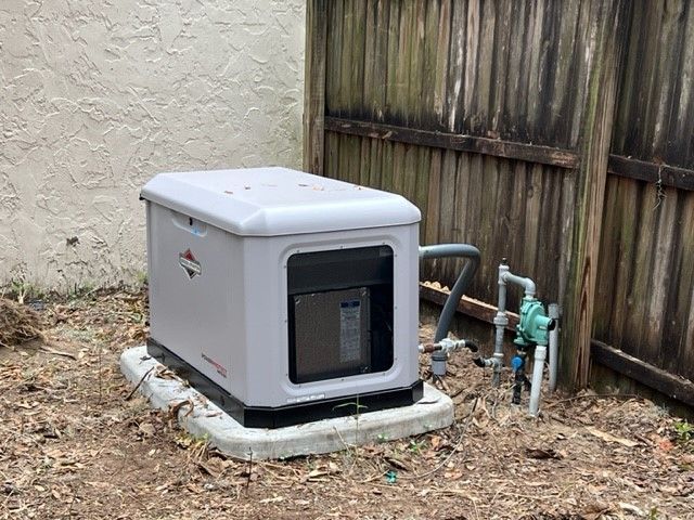 Gray generator with control panel, next to a gas valve near a wooden fence and building.