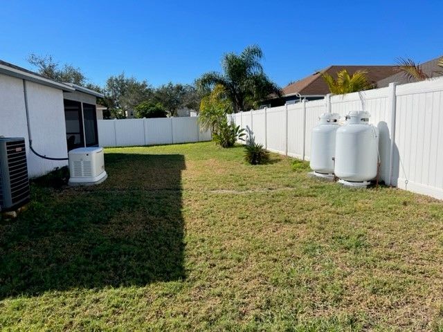 Lawn with white fence, two propane tanks, and an AC unit next to a house under a clear blue sky.