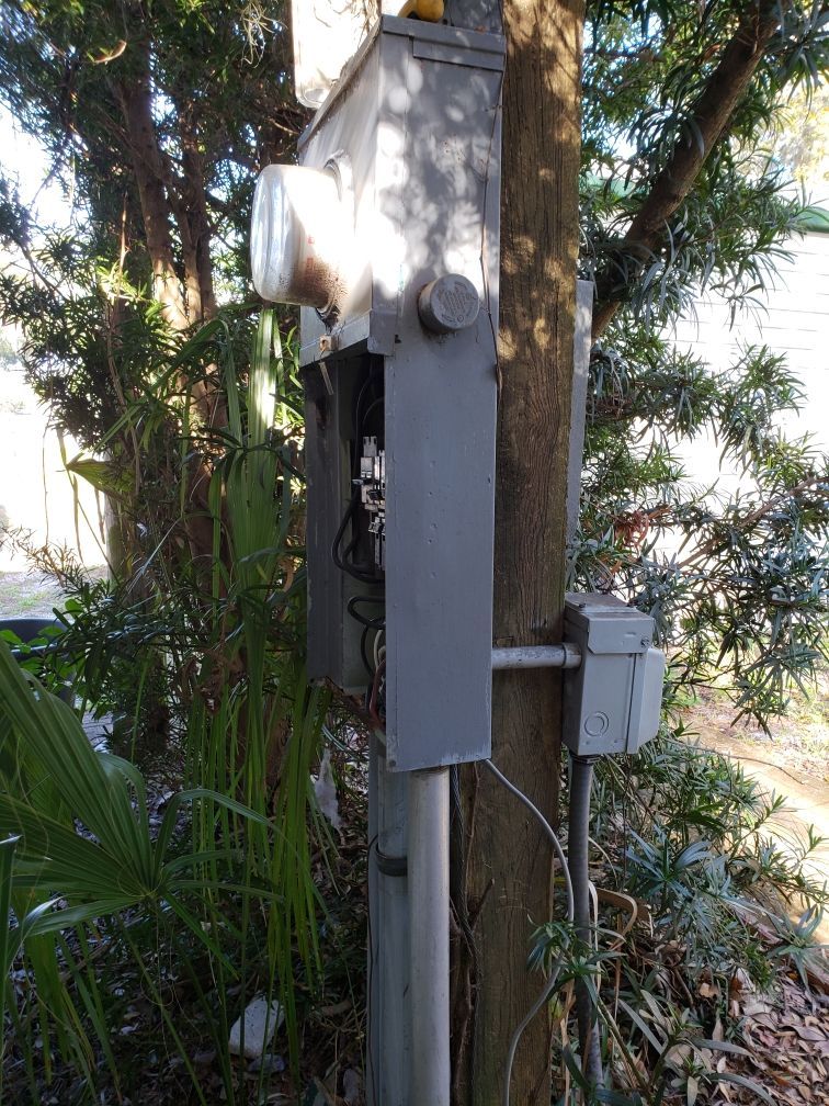 Electric meter box on a pole, mounted near trees and foliage. Gray box with visible wires.