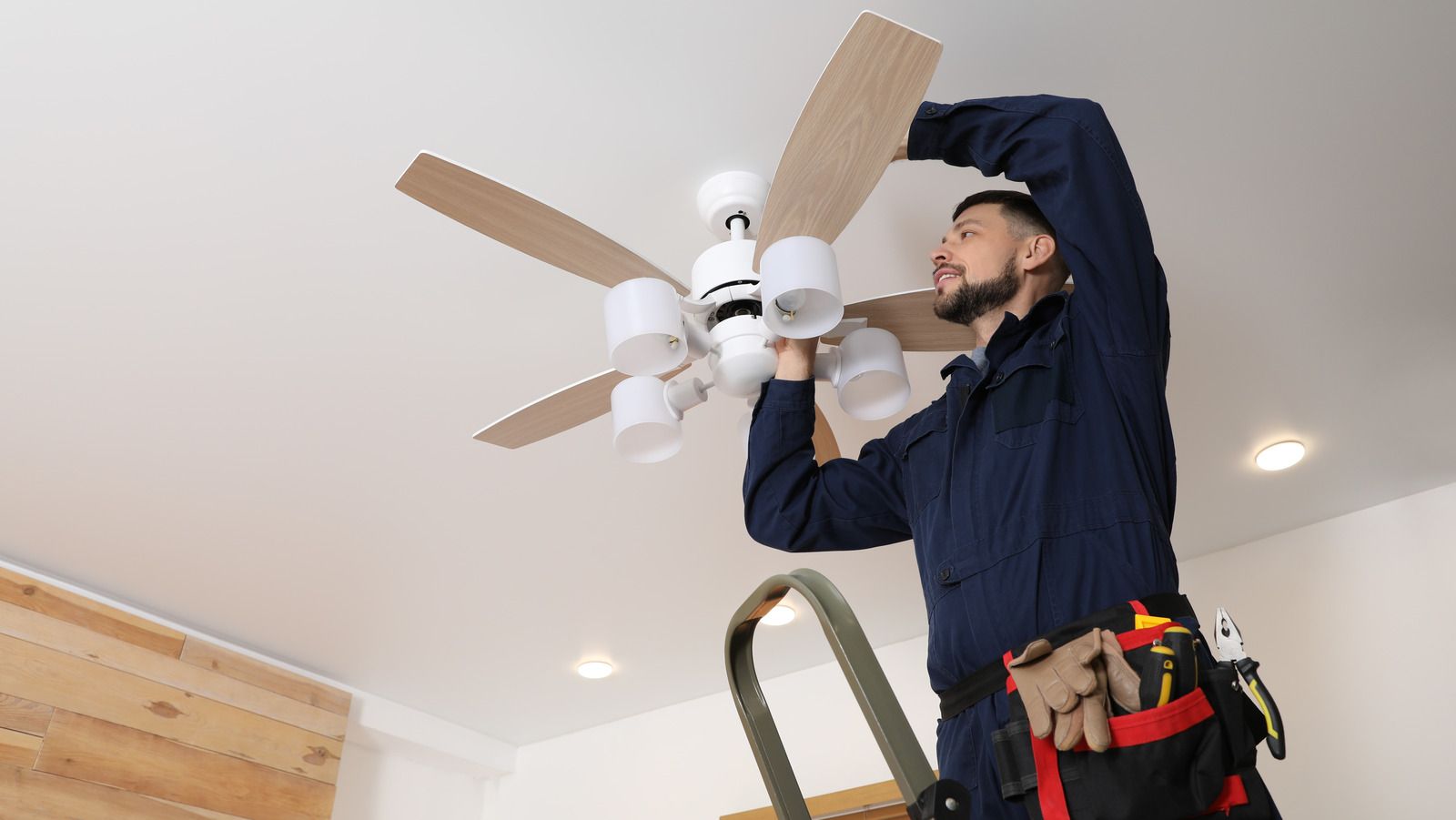 Man installing a ceiling fan, standing on a ladder. He's wearing a blue jumpsuit.