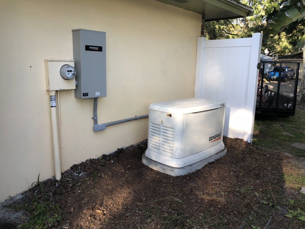Generator and electrical panel mounted outside a light-colored building; a white fence is to the right.