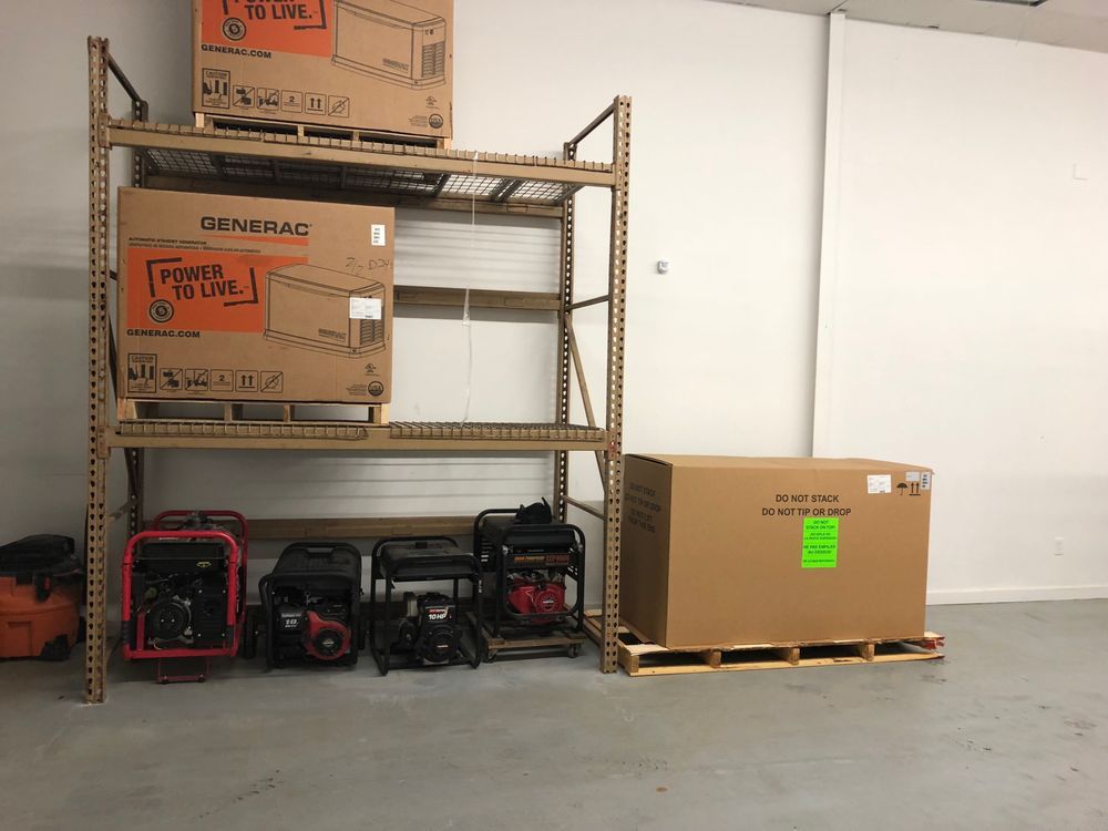 Generators in a warehouse, some boxed, stacked on a metal shelf, others on the floor with a large box on a pallet.