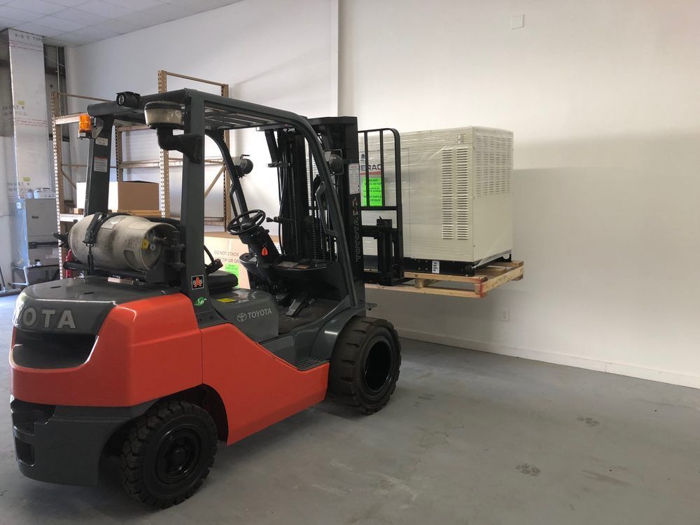 Forklift with an orange and gray body carrying a pallet with a large white container against a white wall.