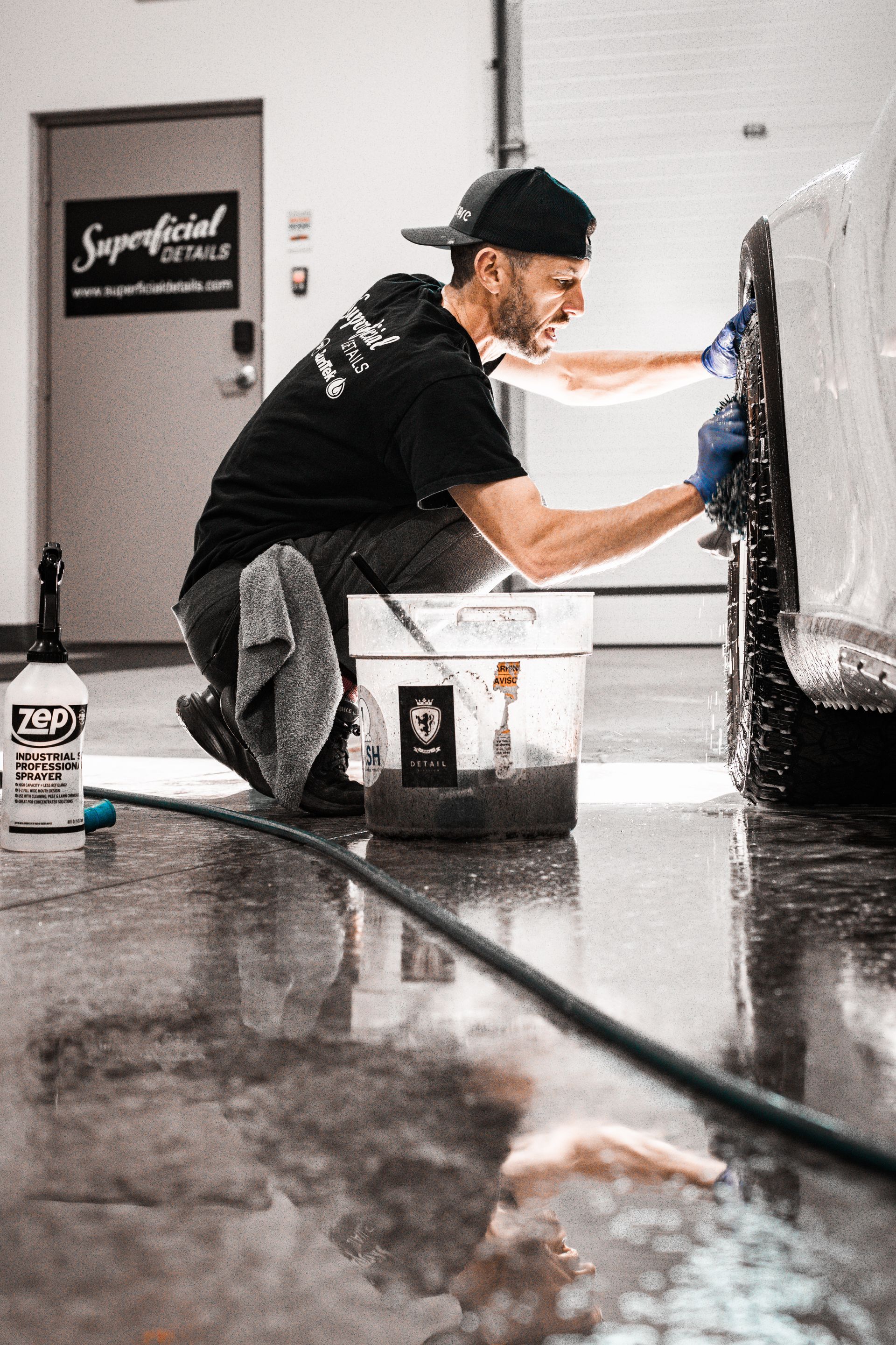 Man washing a car in a garage, wearing a hat and gloves, using a bucket of water.