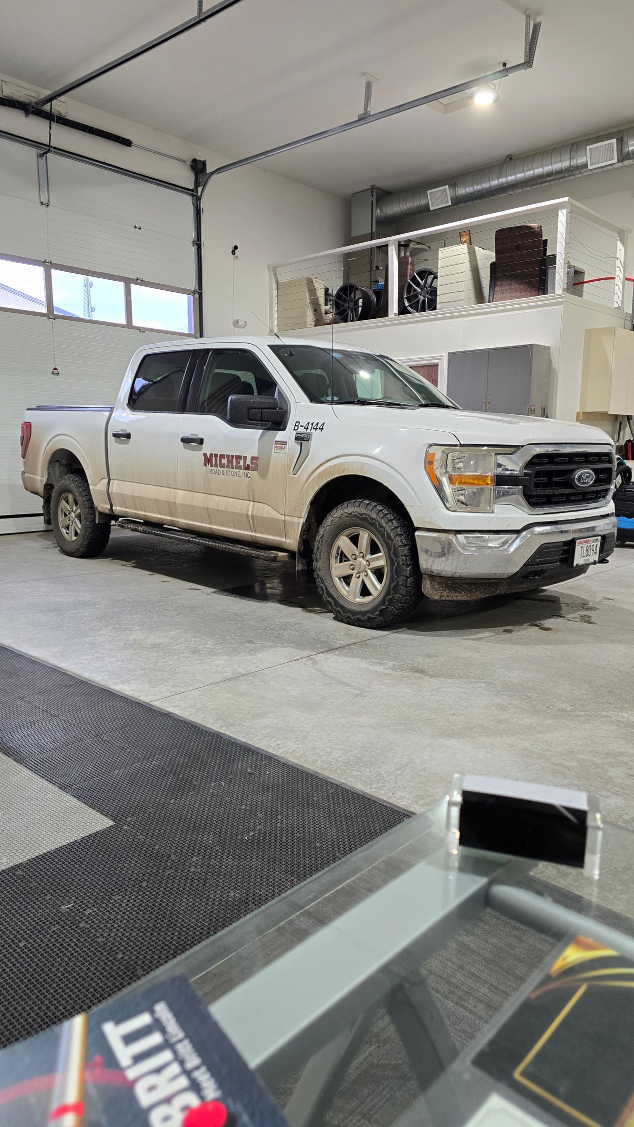 A white Ford F-150 truck covered in mud, parked inside a brightly lit garage.