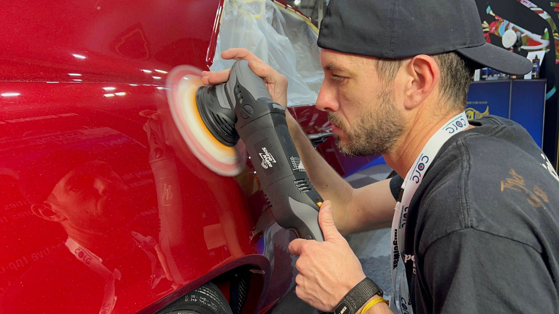 Man using a polisher on a red car panel. He wears a black cap and concentrates on the work.