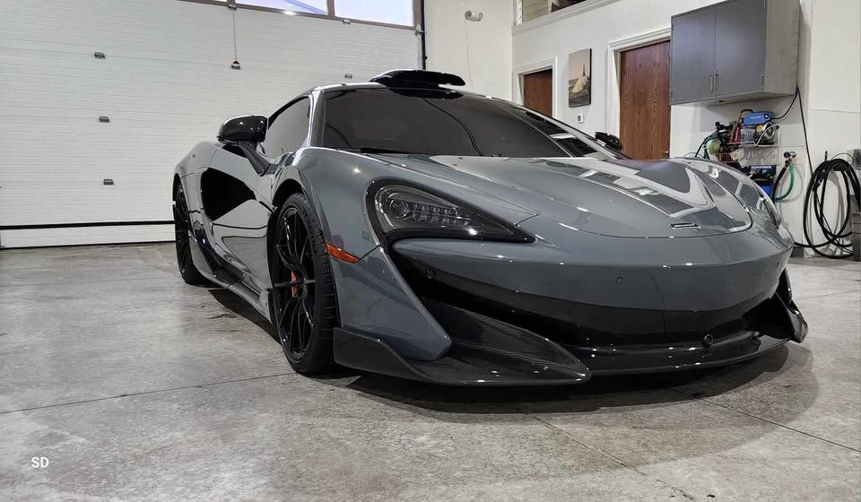 Gray McLaren sports car parked inside a garage with open door and equipment on wall.