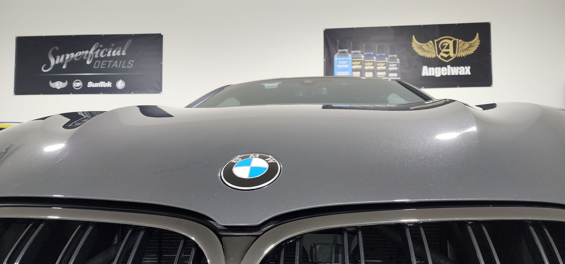 A black BMW car's front with the logo is in a detailing studio, with two signboards in the background.