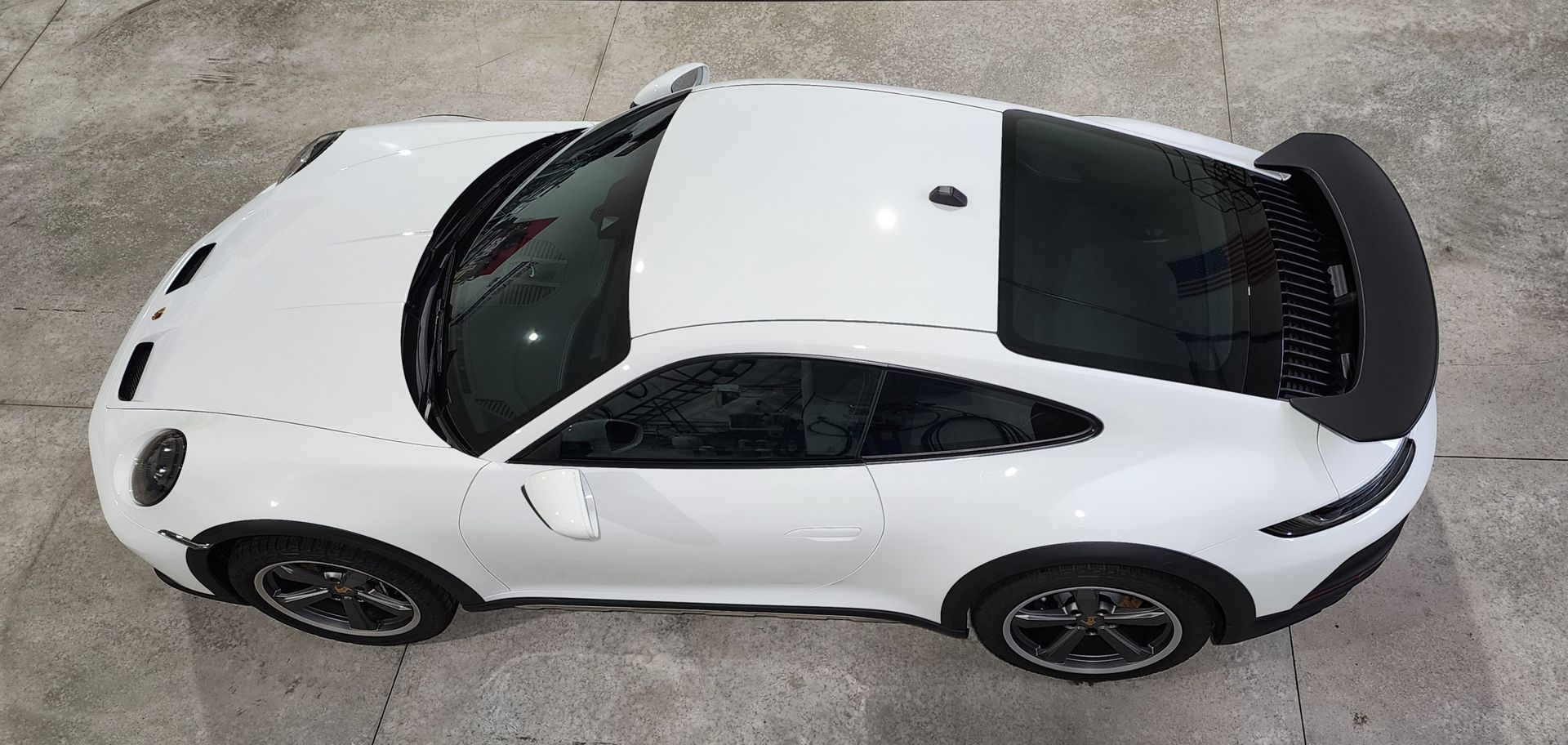 A white Porsche 911 coupe viewed from a high-angle, overhead perspective parked on a concrete surface.