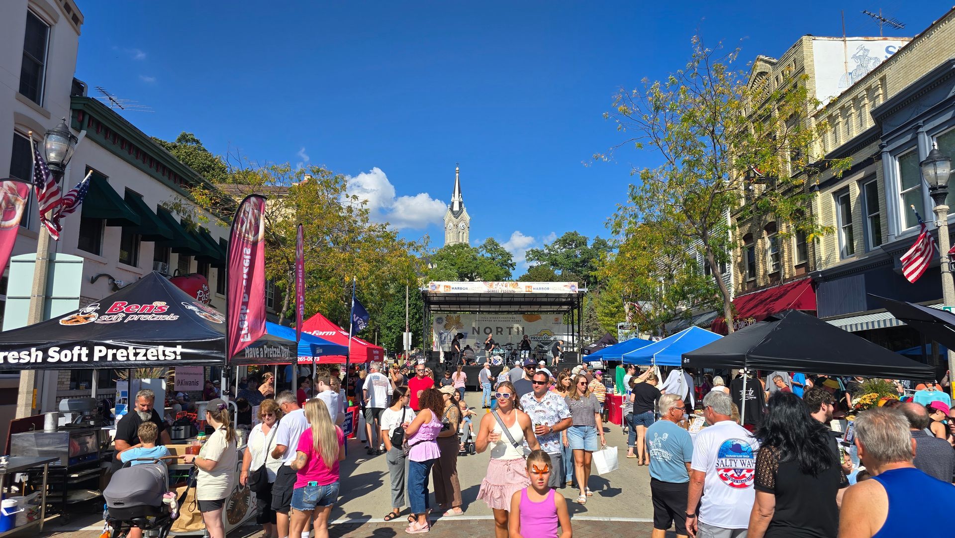 Crowd at an outdoor festival on a sunny day. Tents line the street with a stage in the background.