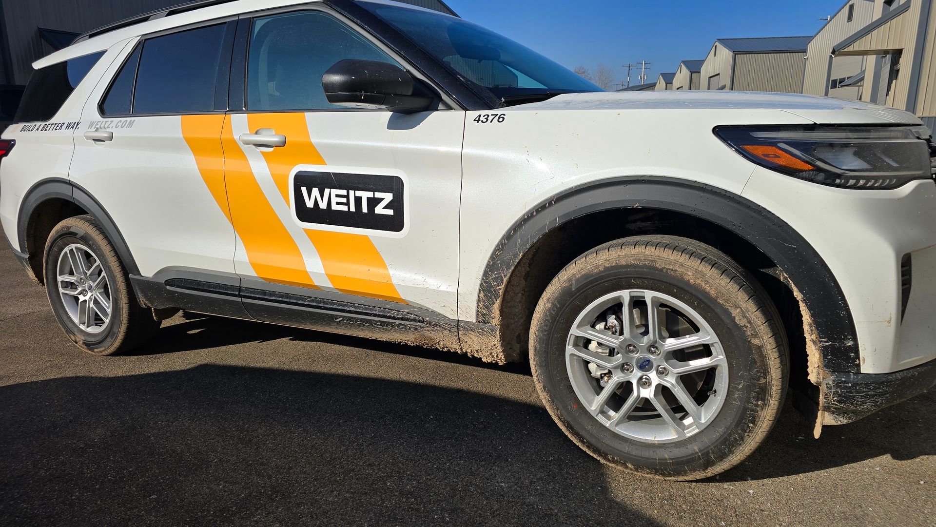 A white Weitz branded SUV with orange stripes on the side, parked on a dirt lot.