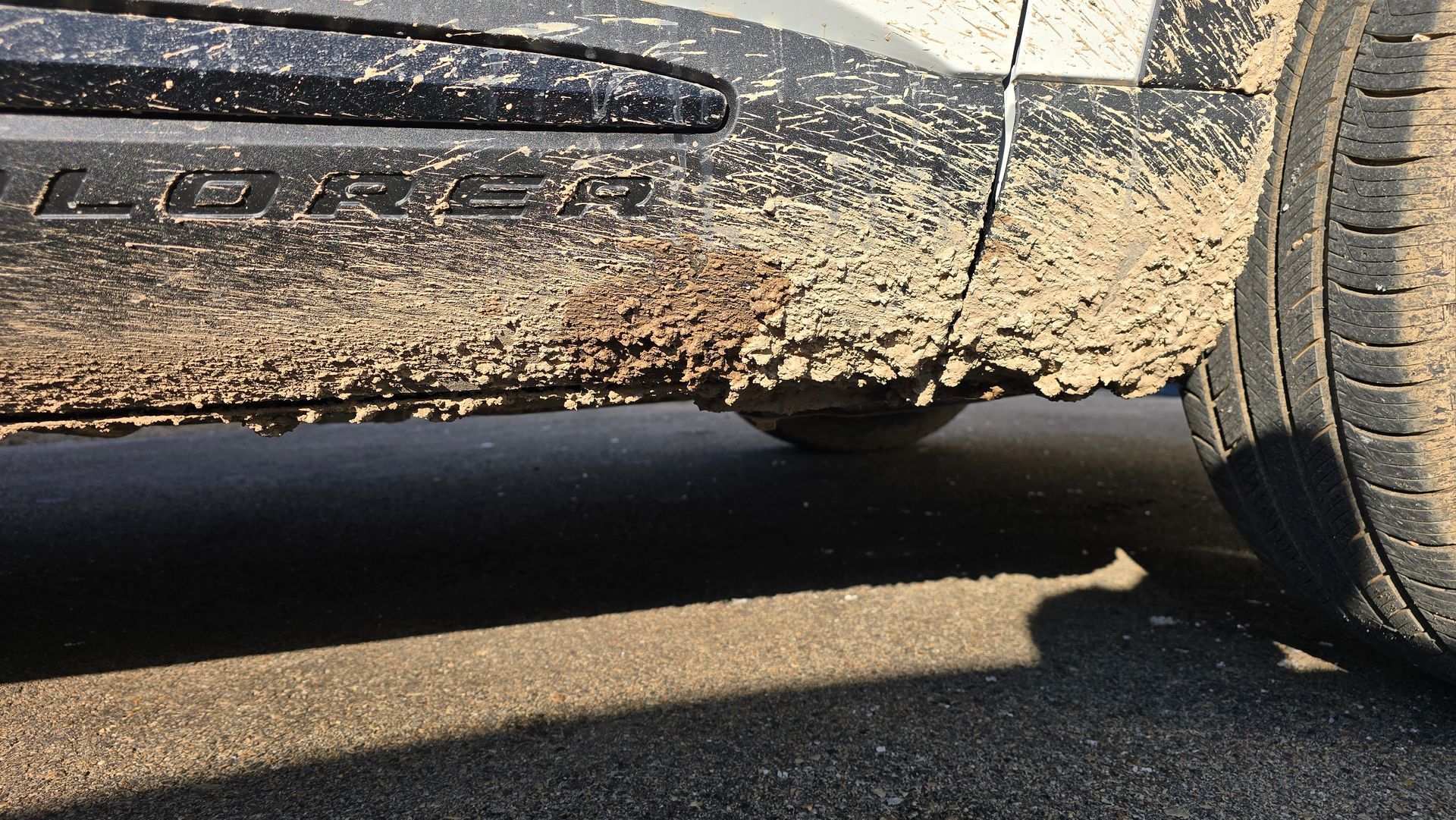 Close-up of a muddy car door panel and tire on a grey asphalt surface.