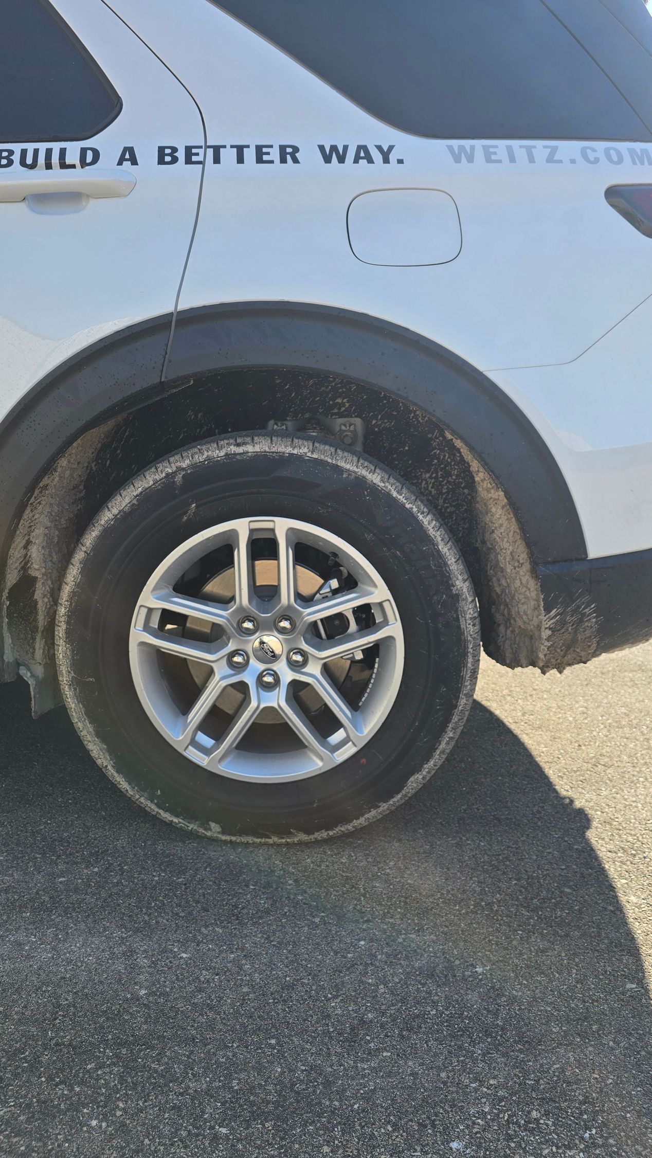A close-up of a white SUV's rear tire, which is covered in dirt, with a partial company logo visible on the panel above.
