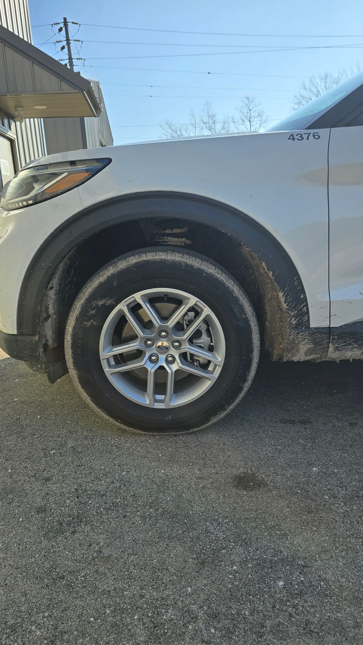 Side view of a white vehicle's front wheel and fender, parked on a gravel surface under a clear sky.