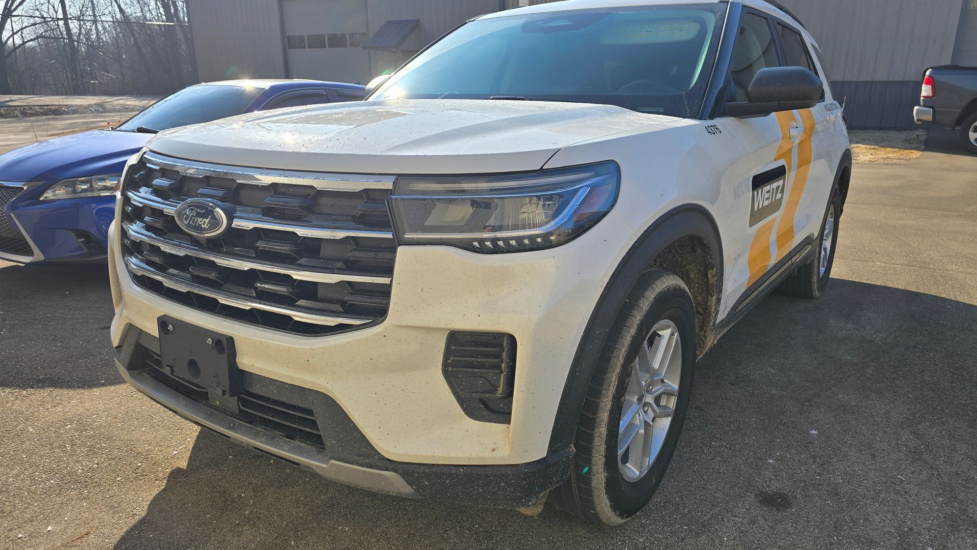 A white Ford Explorer SUV parked on a gravel lot with a partial view of a blue vehicle behind it.