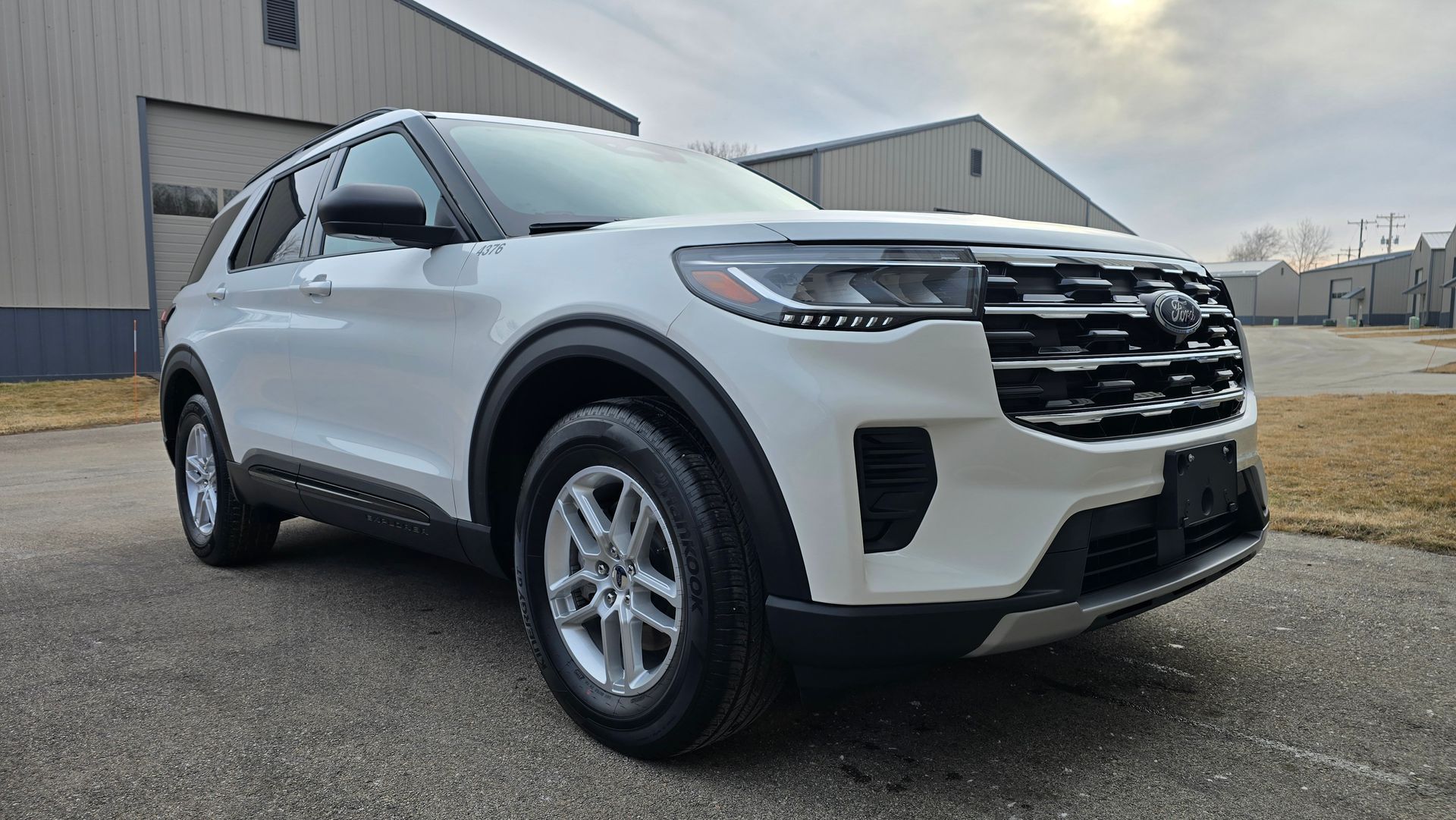 A white Ford Explorer SUV parked on an asphalt lot in front of two metal industrial buildings under a cloudy sky.