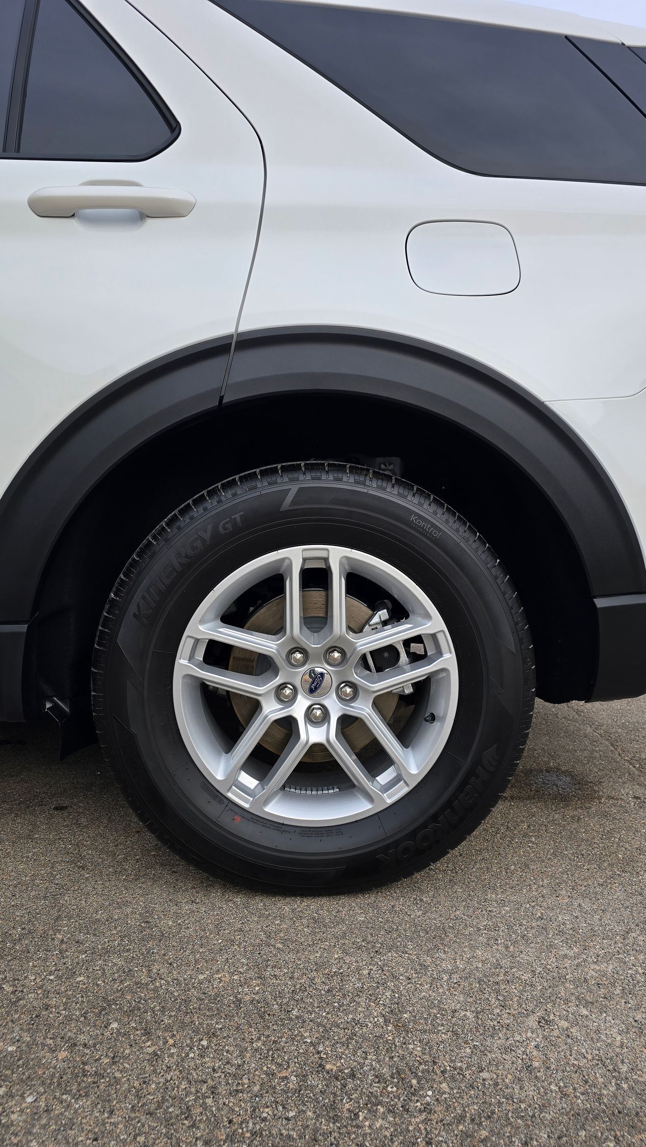 A close-up of a white SUV's rear tire and fender, featuring a silver multi-spoke wheel and black plastic trim.