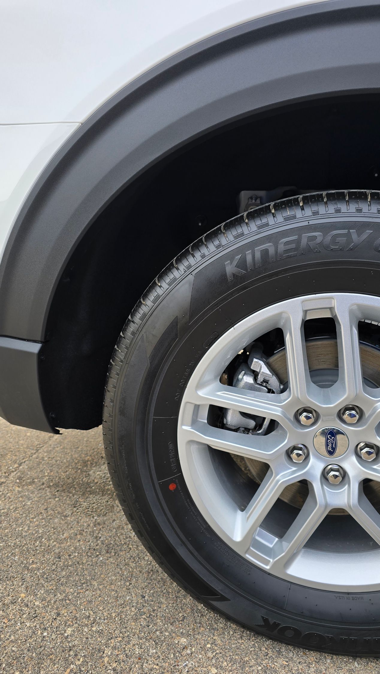 A close-up view of a car's silver alloy wheel and tire with a black plastic wheel arch trim against a gravel background.
