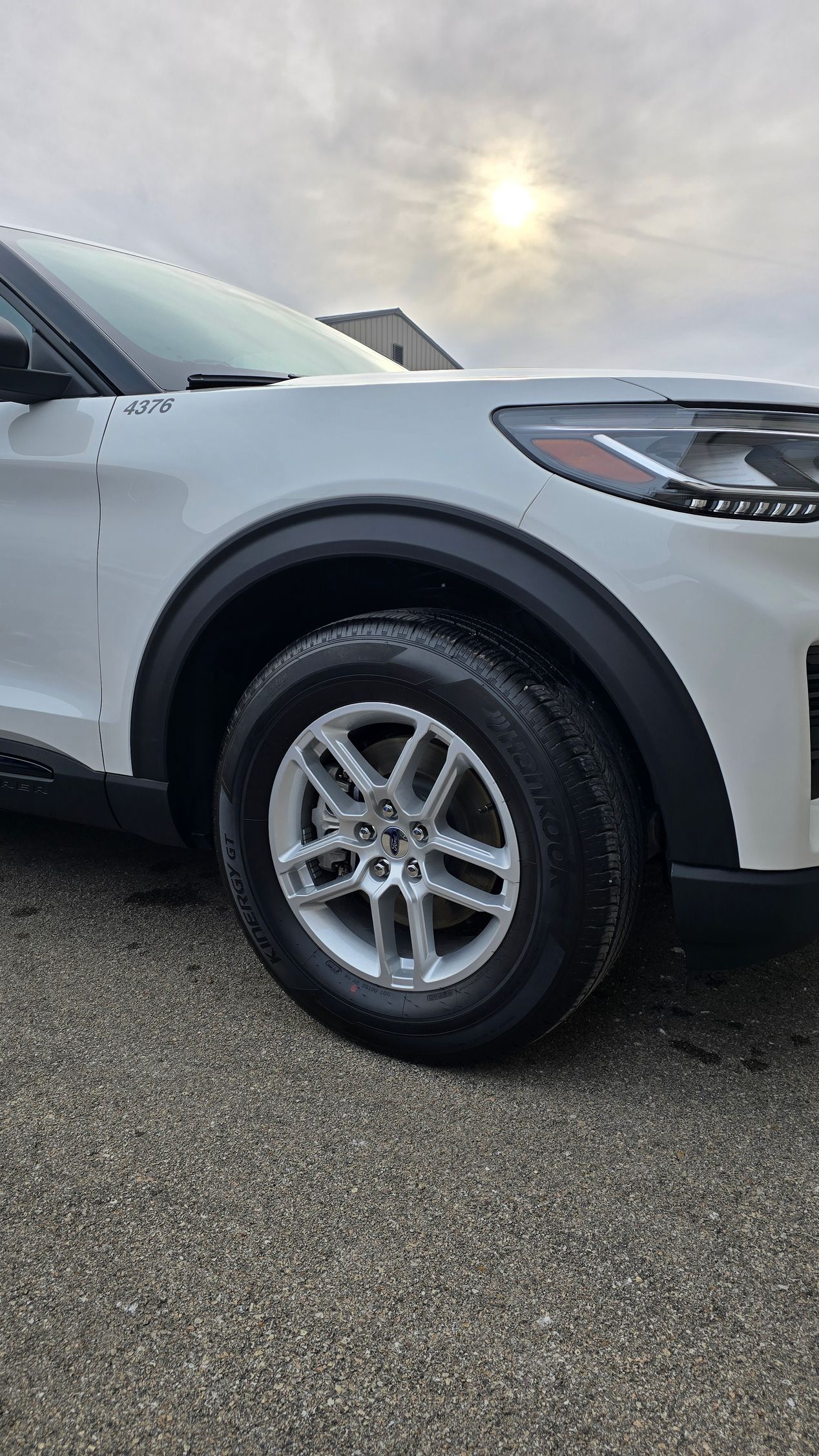 Close-up of a white SUV's front wheel and fender, parked on a gravel surface under a bright, cloudy sky.