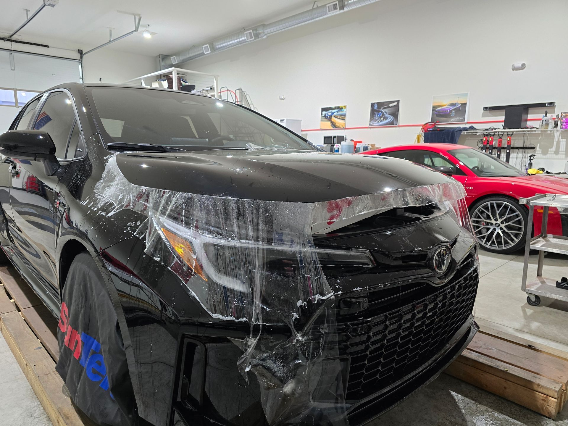 Black car with protective film applied, inside a shop. A red car is in the background.