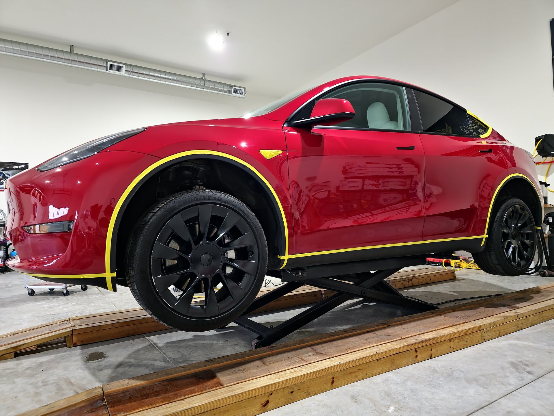 A red Tesla Model Y on a scissor lift in a garage, with yellow masking tape applied along its wheel wells and side panels.