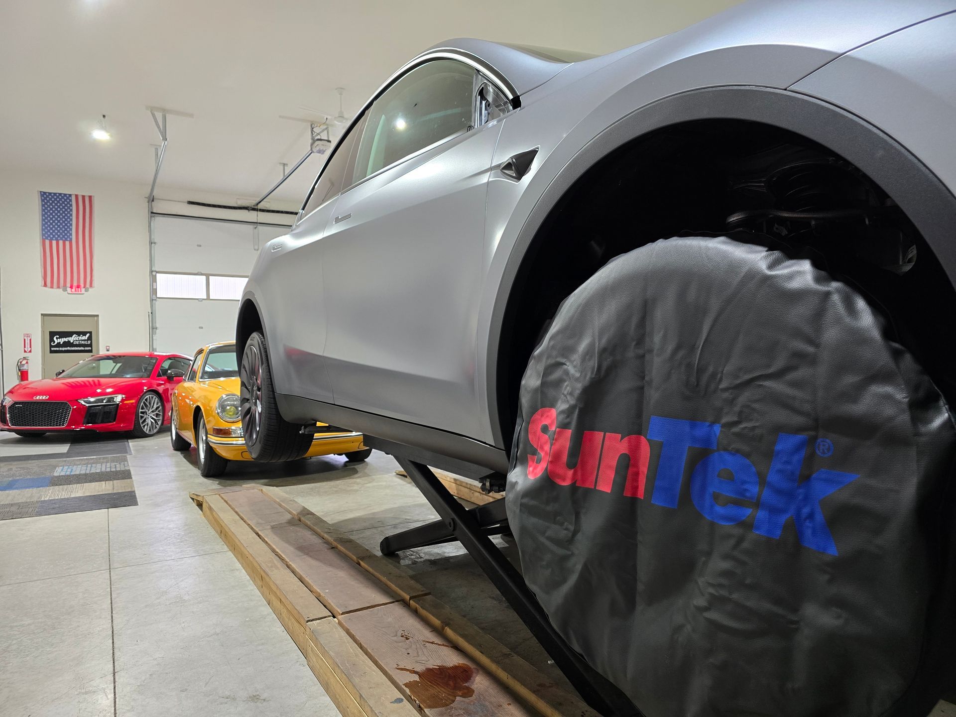 A silver Tesla raised on a lift in a garage, wheel covered, with a red car and yellow car visible in the background.