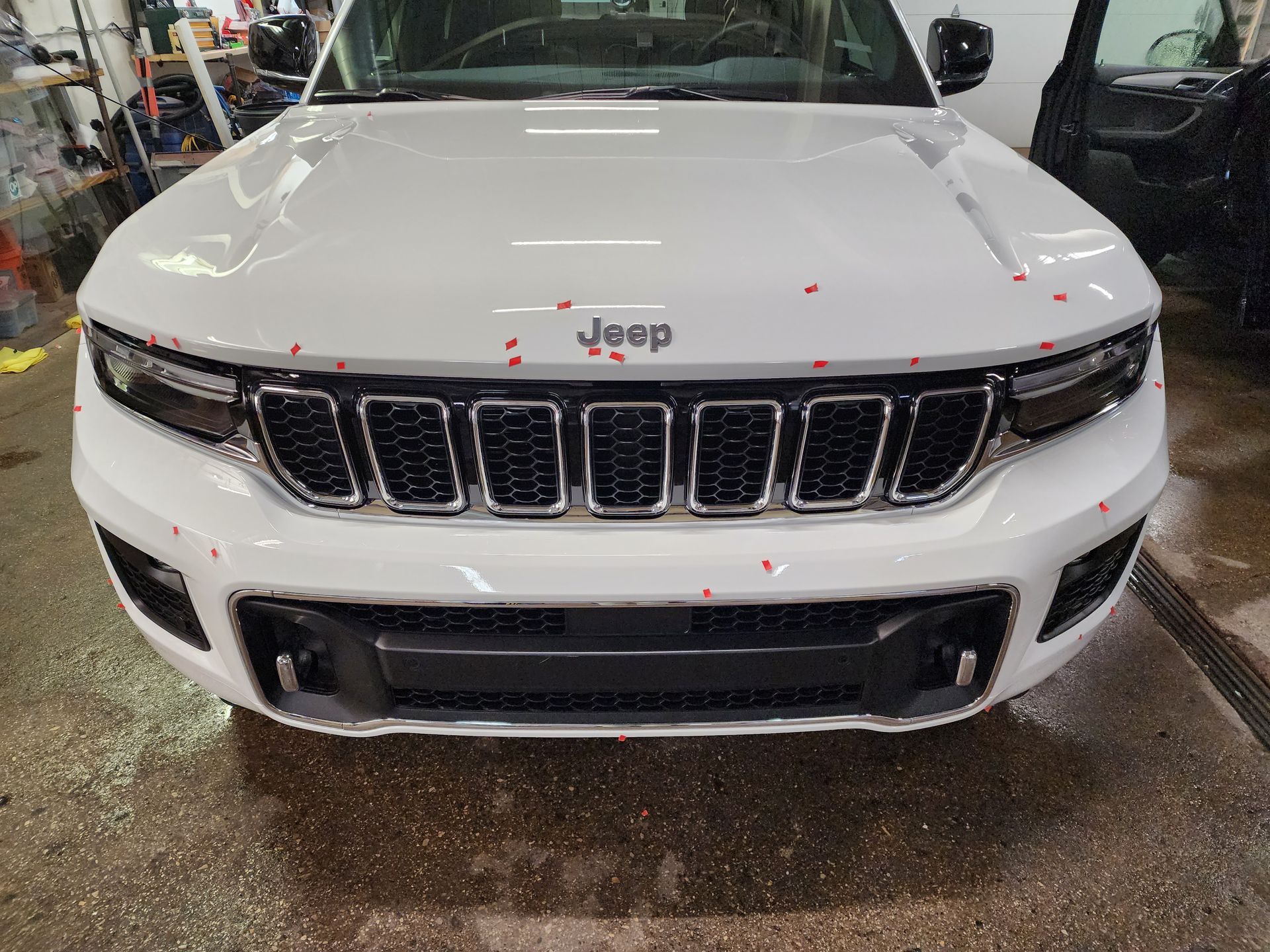 Front view of a white Jeep Grand Cherokee parked inside an automotive workshop with red markers on the body panels.