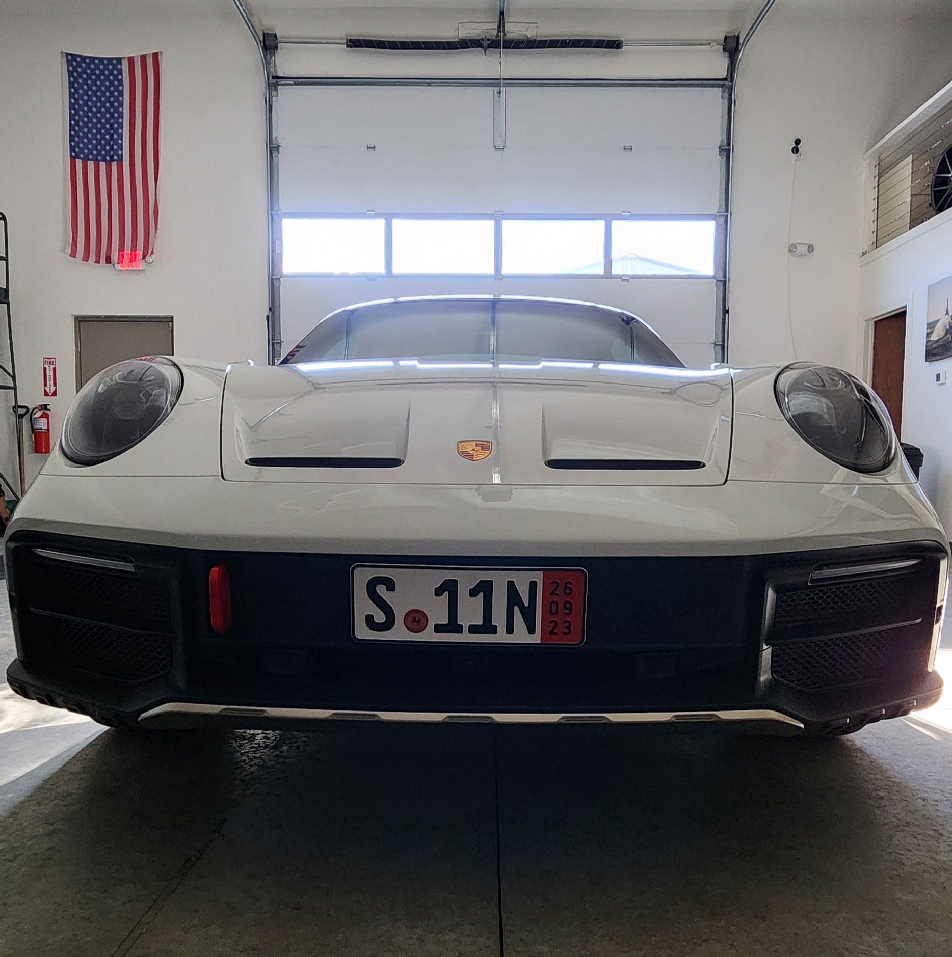 A white Porsche parked in a garage with an American flag on the wall and a custom license plate reading S.11N.