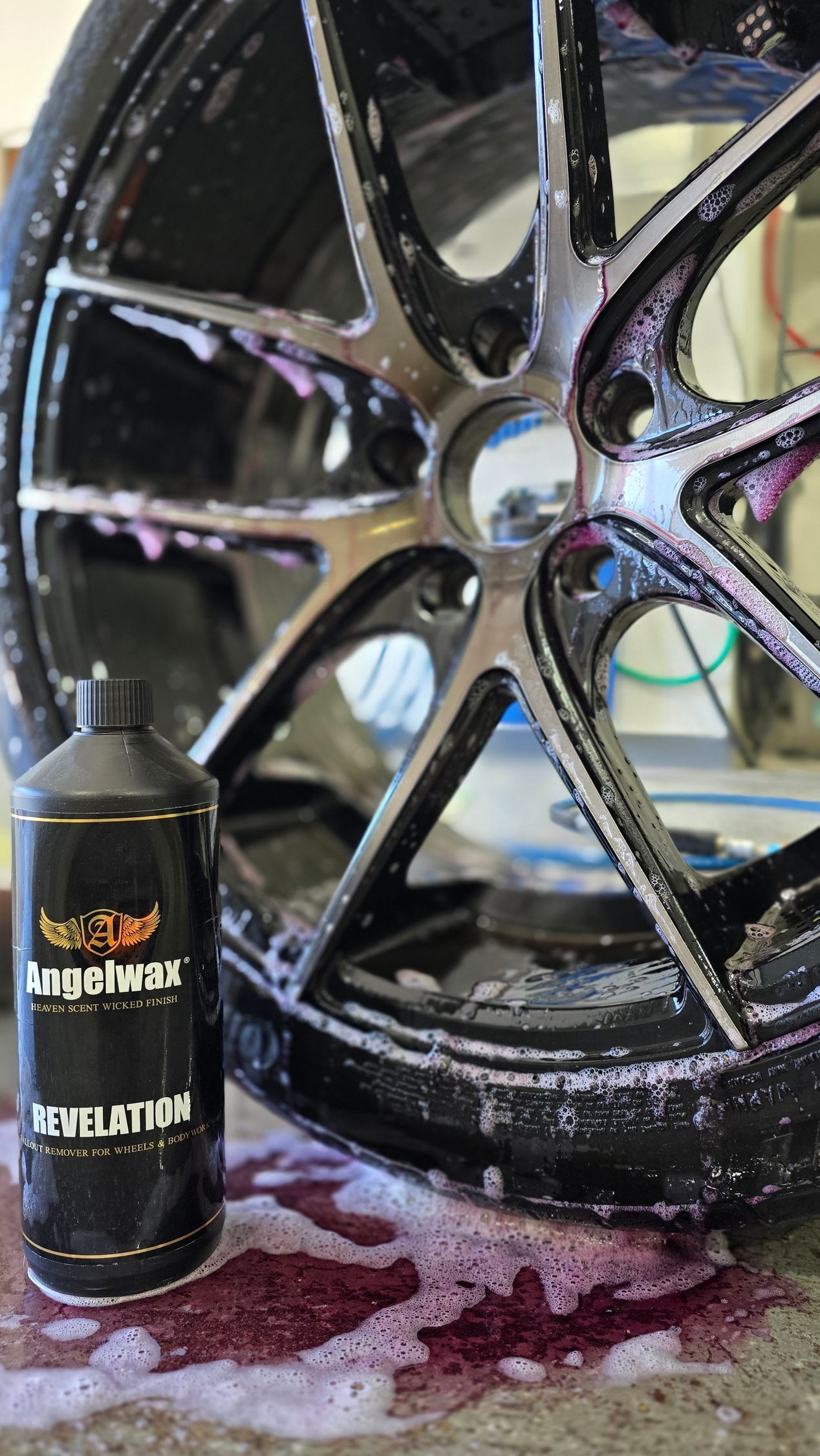 A black car wheel covered in foamy cleaner with a bottle of wheel cleaner next to it.