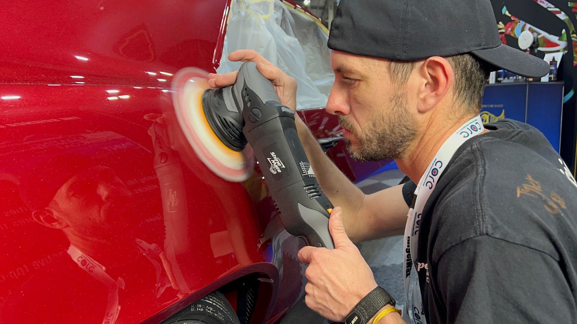 Man polishing a red car with a power buffer. Indoors, wearing a cap and black t-shirt, focused expression.