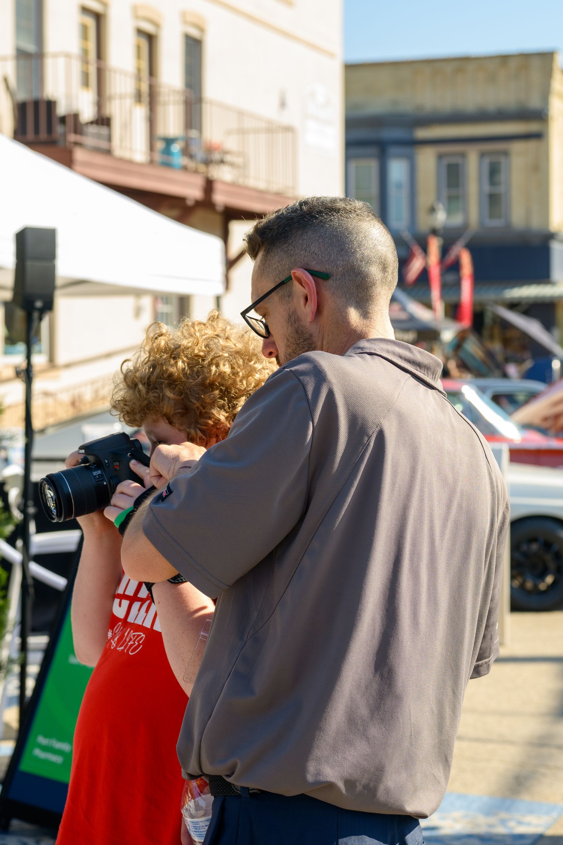 Man and child looking at camera screen outdoors; white building background.