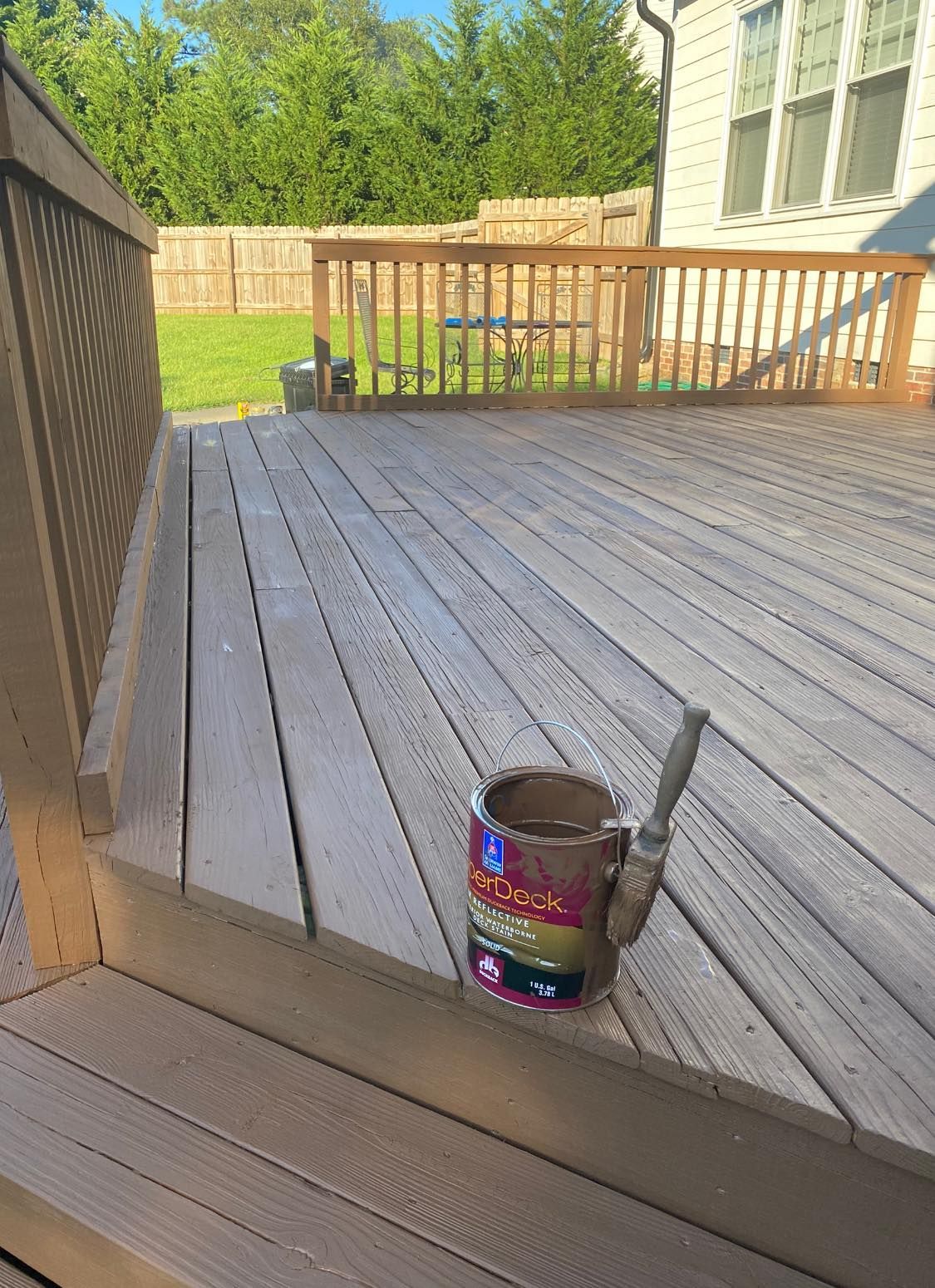 Paint can with a brush on a wooden deck, with a yard and house in the background.