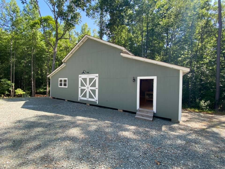 Gray outbuilding with white trim, barn doors, and open entry on a gravel lot surrounded by trees