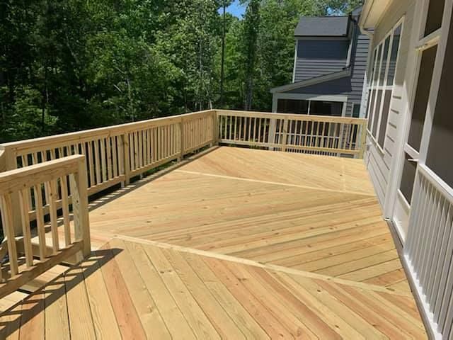 Wooden deck with railing beside a house, overlooking green trees in sunlight