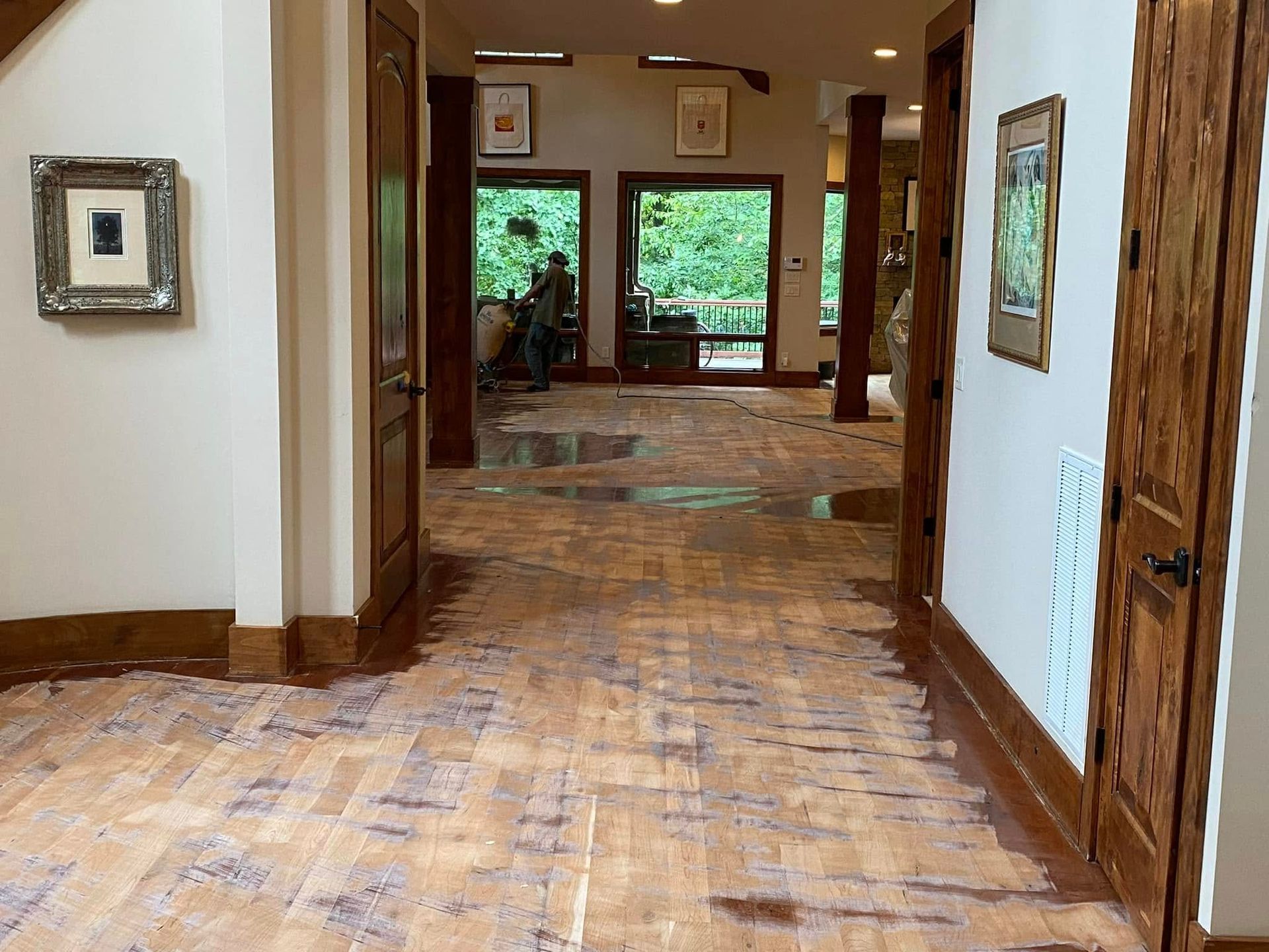 Empty hallway with wood floors, white walls, and large windows showing green trees beyond.